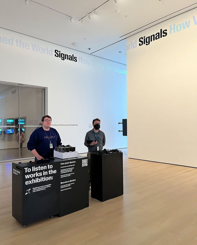 A photo of the entrance to the "Signals" exhibit where two museum attendants hand out smartphones equipped with the Sennheiser MobileConnect app.